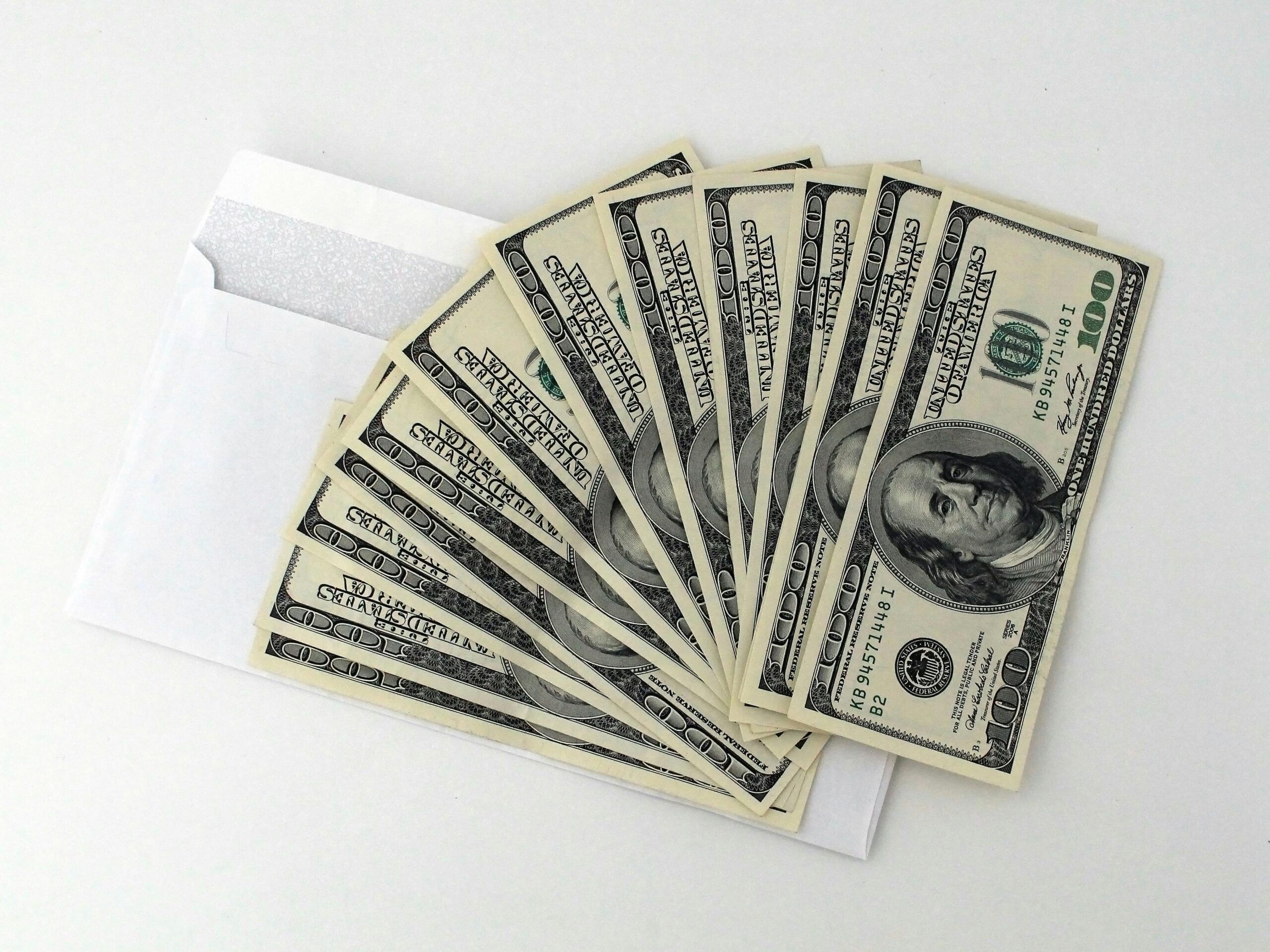 Fan of US $100 bills partially out of a white envelope on a white background.